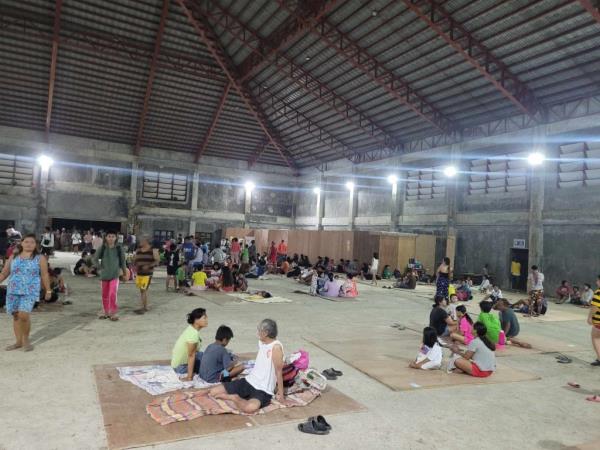 Filipino people cluster in small groups, some standing and some sitting on the floor, in a cavernous warehouse used as an evacuation center after the earthquake.